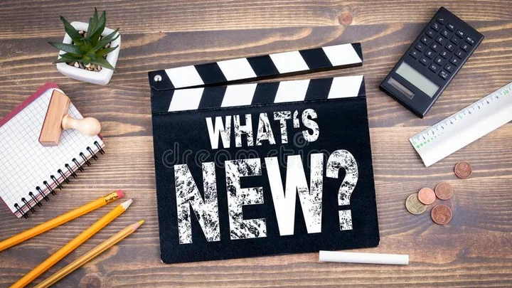 Clapperboard reading ‘What’s New?’ on wooden desk with pencils, notebook, calculator, ruler, coins, and plant.